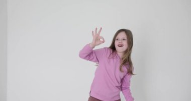 Sevimli küçük kız gülümser ve beyaz arka planda parmakları olan bir işaret gösterir. Uzun saçlı çocuğun gözleri mutluluk ve kendine güvenle parıldıyor.