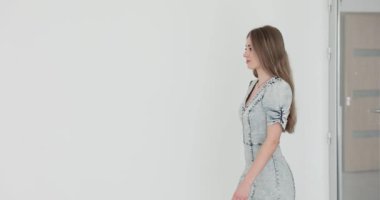 Kendine güvenen, kot elbiseli genç bir kadın beyaz duvara yaslanıyor. Uzun, dağınık saçlı şık bir kadın modeli atölyedeki yeni kıyafetleri gösteriyor.