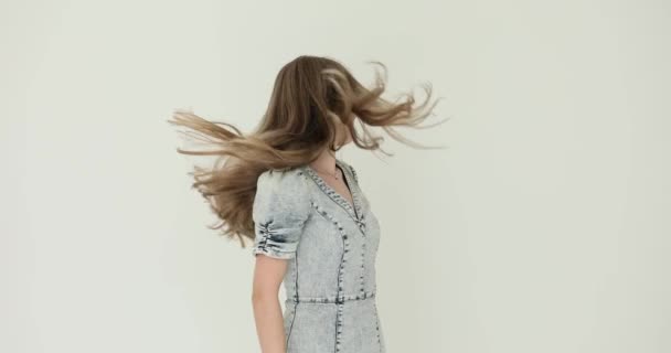 Joyeux jeune femme en robe de denim secoue la tête en jetant de longs cheveux lâches côte à côte sur fond blanc. Belle dame démontre coiffure élégante