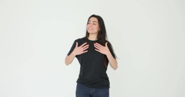 Siyah tişörtlü genç kadın beyaz arka planda minnettarlığını ifade ediyor. Esmer kadın ellerini göğsüne koyup gülümseyerek konuşuyor.