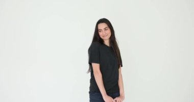 Uzun, bol saçlı, gülümseyen ve beyaz arka planda çenesine dokunan genç bir kadın. Siyah tişört ve kot giyen kadın model.