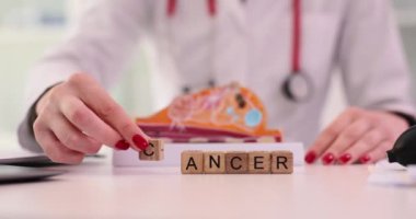 Doktor kadın eli, tahta küpün üzerine mektup ekliyor. Masadaki yapay göğüs modeline karşı Kanser kelimesi yazıyor. Uzman kadın organlarında ciddi bir hastalık olduğunu söylüyor.
