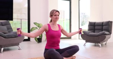 Sportif kadın, modern apartman dairesinde paspasın üzerinde oturan dambıllarla el kaldırıyor. Vücudu güçlendirmek ve ev yapımı ağır çekimde sağlığı korumak