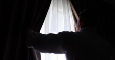 Man in white bathrobe opens heavy curtains covering panoramic window to illuminate room. Male person admires view from window in early morning