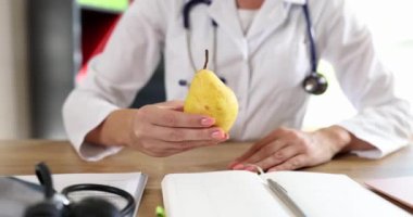 Female doctor gives patient fresh pear sitting at table in clinic office. Nutritionist in medical uniform shows organic fruit. Healthy diet concept slow motion