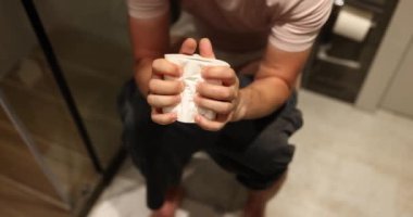 Male person grips toilet paper tightly sitting on toilet bowl in apartment bathroom. Concept of daily routine in modern premise slow motion