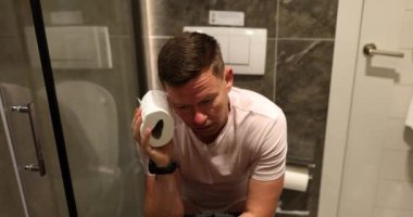 Man applies toilet paper to head sitting on toilet bowl near shower. Concept of daily routine in light bathroom of accommodation slow motion