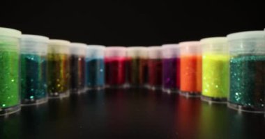 Sealed jars with multi-colored shiny glitter stand in circle in dark studio. Vials with sequins on black surface in premise. Bright decoration designed for makeup or manicure