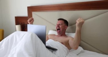 Male freelancer rejoices looking at laptop monitor on bed with white blanket at home. Concept of good deal and online work in comfortable conditions slow motion