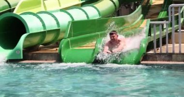 Cheerful male person in swimming trunks descends from water slide into pool in water park. Entertainment and recreation concept at resort slow motion
