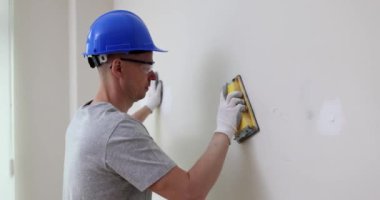 Builder in helmet and goggles polishes plaster spot on wall in restored room. Employee fixes holes and trenches on drywall panel with tool