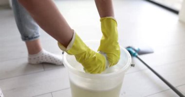 Woman in rubber gloves wrings foamy mop rag over bucket with water in room. Person works with floor house cleaning supplies slow motion