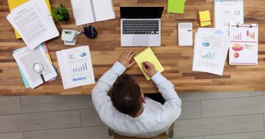 Businessman writes list of future tasks sitting at wooden desk with laptop and papers. Manager schedules work at workplace in office upper view