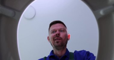Funny plumber opens toilet bowl and scratches nape with plunger in restroom. Emotional man cleans clog in pipe with traditional tool view from bottom