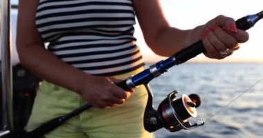 Woman in striped tank top fishes with modern spinning on boat slow motion. Fisher woman with rod pulls out marine catch while sailing at sunset