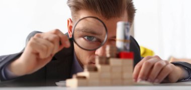 Close-up of businessman holds in hand black magnifying glass looks at tower of coins placed on wooden cubes. Financial development, invest, growth concept clipart