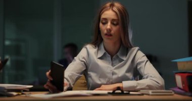 Yorgun bir kadın telefonda zamanı kontrol eder ve kafasını geç saatlere kadar masaya koyar. Ofis atmosferi, son teslim tarihinin yaklaşmasından kaynaklanan stres ve yorgunluğu yansıtıyor.