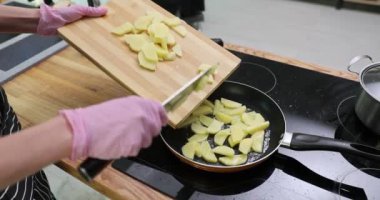 Pembe eldivenli aşçı bıçakla tahtayı kesmekten dilimlenmiş patatesleri kızgın tavaya ekler. Yemekçi akşam yemeği için yemek pişirmeye başlar.