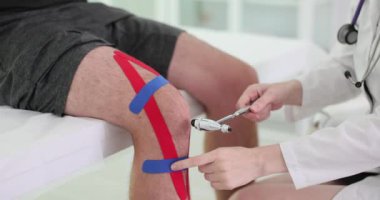 Doktor refleks çekiçle dizini kontrol ederek sinir tepkisini test ediyor. Kineziyoloji bandı olan hasta özel klinikte muayene ediliyor.