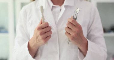 Diş hekiminin ellerinde diş fırçası ve diş aletleri var. Üniformalı kadın doktor, hastalıkları önlemek için ağız temizliğine ihtiyaç olduğunu açıklıyor.
