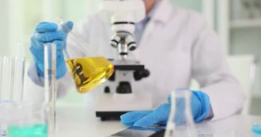 Eldivenli bilim adamı, Erlenmeyer matarasını iş yerinde sarı sıvıyla döndürüyor. Laboratuvar çalışanı üniforma analizi laboratuarda yağlı çözelti