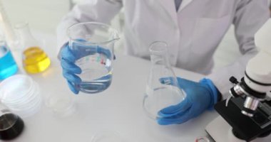 Bilim adamı laboratuardaki iş yerinde şişeden mataraya su döküyor. Lateks eldivenli işçiler kimyasal araştırmalar için malzeme hazırlarlar.