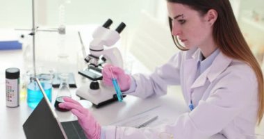 Kadın bilim adamı elinde turkuaz sıvıyla cam tüp tutuyor ve tablete veri yerleştiriyor. Üniformalı kadın laboratuvar çalışanı araştırmadan sonra rapor hazırlıyor