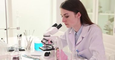 Üniformalı kadın bilim adamı laboratuvarda mikroskop kullanarak araştırma yapıyor. Eldivenler üzerinde odaklanmış kadın çalışanlar örnekleri incelikle analiz eder.