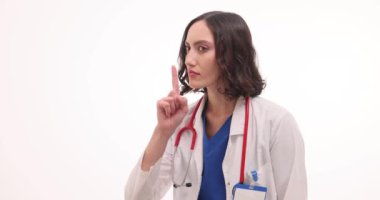Kadın doktor, beyaz arka planda sessizlik jesti yaparken parmaklarını ağıza koyuyor. Steteskoplu kadın doktor hastaya randevuda sessiz olmasını söylüyor.