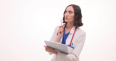 Üniformalı düşünceli kadın doktor elinde bir not defteri tutuyor ve beyaz arka plan üzerine notlar yazıyor. Ciddi yüz ifadesi olan doktor hasta verilerini kaydediyor.