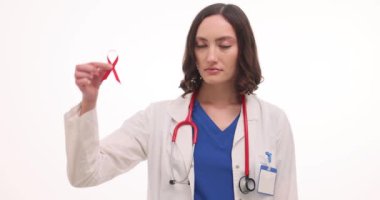 Kadın doktor beyaz arka planda kırmızı kurdele ve prezervatif tutuyor. Üniformalı ciddi kadın sıhhiyeci HIV AIDS bilgisinin önemini hatırlatıyor