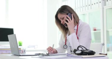 Üniformalı kadın doktor hastanede iş yerinde telefonla konuşuyor. Meşgul sarışın doktor, dikkat çeken hastalarını dinler.