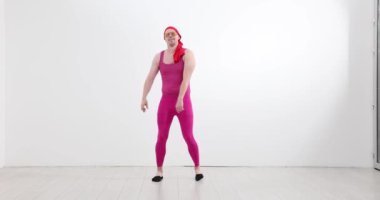 Hevesli erkek fitness eğitmeni pembe kıyafetler giyiyor ve beyaz duvara karşı bantla dans ediyor. Dansçı enerjik aerobik yapıyor.