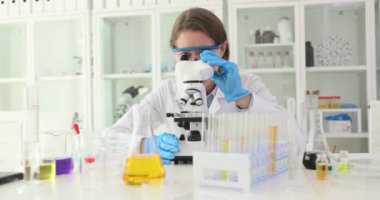Güvenlik gözlüklü pozitif kadın bilim adamı laboratuvarda mikroskopla çalışıyor. Kadın kimyager, temiz eczane laboratuvarında biyolojik materyalleri analiz ediyor.