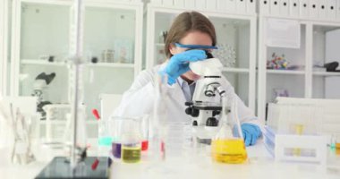 Bornoz ve güvenlik gözlüğü takan kadın bilim adamı laboratuarda mikroskop kullanıyor. Dost canlısı bayan eczacı kameraya el sallayarak testler yapıyor.