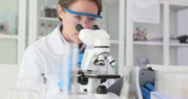 Kadın araştırmacı örnekleri mikroskop altında inceleyip laboratuardaki panoya not alıyor. Bilim adamı laboratuarda numune incelemesi yapıyor.