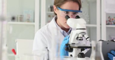 Koruyucu gözlüklü kadın biyokimyacı laboratuvar ortamında örnekleri mikroskop altında analiz ediyor. Odaklanmış kimyager laboratuvarda materyal araştırıyor.