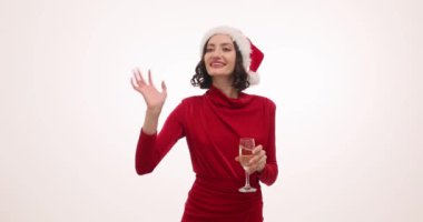 Noel Baba şapkalı ve kırmızı elbiseli heyecanlı kadın beyaz arka planda şampanya kadehiyle el sallıyor. Güzel bayan gülümsüyor Noel 'de mutlu görünüyor