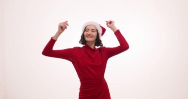 Noel Baba şapkalı neşeli genç bayan beyaz arka planda neşeyle dans ediyor. Bayanın gülümsemesi Noel partisinde mutluluk saçıyor ağır çekimde