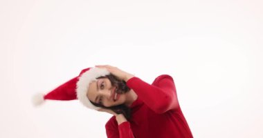 Kıvırcık saçlı güzel bir kadın Noel Baba şapkasını beyaz arka planda düzeltirken güler. Olumlu ifadeli kadın tatil ruhu yayıyor.