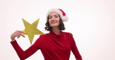 Noel Baba şapkası takan mutlu kadın beyaz arka planda altın yıldızlar gösteriyor. Terracotta kazaklı, gülümseyen genç bir kadın partideki neşeli atmosferden hoşlanıyor.