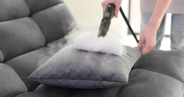 Ev hanımı evdeki kanepede buharlı temizleme yastığı kullanıyor. Önlüklü dikkatli kadın, tekstil nesnelerinden toprağı temizleme işlemine girişir.