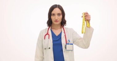 Kadın doktor beyaz arka planda mezura ve çikolata gösteriyor. Beyaz cüppeli kadın beslenme uzmanı başını sallayarak diyeti hatırlatıyor.