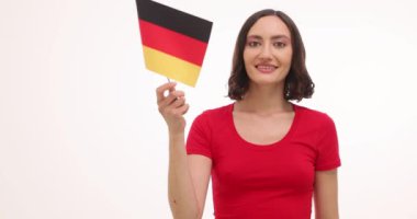 Kırmızı tişörtlü genç kadın beyaz arka planda Alman bayrağı sallıyor. Gülümseyen esmer seyahat acentesi ulusal sembolü kullanarak neşe ve vatanseverlik ifade ediyor.