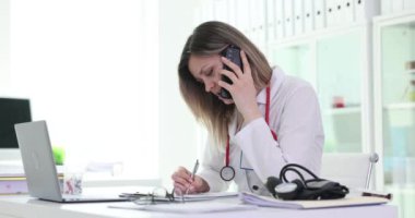 Bayan doktor telefonda konuşurken notlar alır. Meşgul sarışın sağlık uzmanı modern ofis ortamında birden fazla görevi yerine getiriyor.