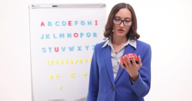 Genç bayan öğretmen kırmızı saate bakıyor ve İngiliz alfabesine karşı sınıfı bekliyor. Dişi pedagog, öğrencilerin yavaş çekimde geç kalmasının nedenlerini düşünüyor