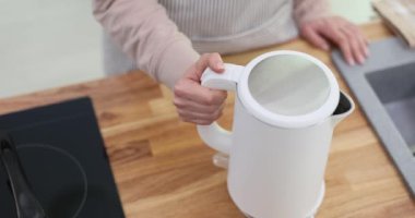 Önlüklü kadın su kaynatmak için modern elektrikli su ısıtıcısını çalıştırıyor. Günlük rutin ev aletleri için şık ve işlevsel