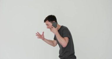 Siyah tişörtlü adam öfkeyle beyaz arka planda telefonla konuşuyor. Erkek oyuncu, ağır çekimde hararetli bir konuşma sırasında hayal kırıklığı yaşıyor