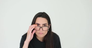 Uzun, gevşek saçlı genç bir kadın beyaz arkaplana yakından bakarak gözlüklerini ayarlıyor. Ciddi yüz ifadesi ve görme sorunu olan bir kadın.