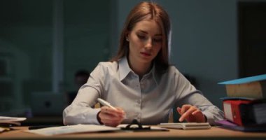 Kadın çalışanlar karanlık ofisteki mali verileri hesaplamaya odaklanırlar. Hesap makineli kadın muhasebeci not defteri sayfasına bilgi yazar